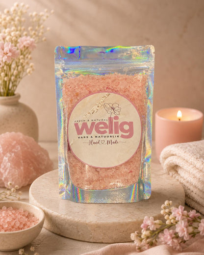 Packaged pink bath salts with a welig label on a decorative setting with flowers and a candle.